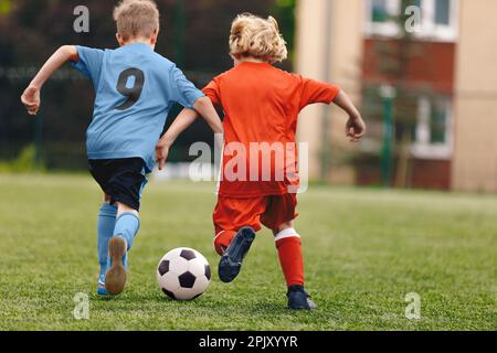 Zwei Schuljungen treten einen Fußball. Schuljungen in roten und blauen Fußballtrikots treten bei einem Fußballspiel gegeneinander an. Fußballduell im Kinderfußballspiel Stockfoto