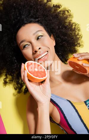 Draufsicht einer fröhlichen afroamerikanischen Frau in bunten Badeanzügen mit Grapefruithälften auf gelbem Hintergrund Stockfoto