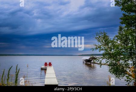 Zwei rote Stühle auf einem Pier mit Blick auf einen See unter stürmischem Himmel in einer Meereslandschaft im Sommer Stockfoto