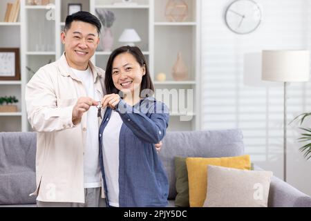 Ein junges, glückliches asiatisches Paar, das lächelt und zusammen in die Kamera schaut, Schlüssel für ihr neues Apartment-Haus hält, Porträt von glücklichen Hausbesitzern. Stockfoto