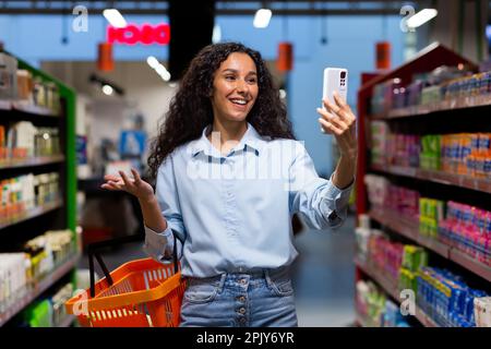 Eine junge lateinamerikanische Frau, Bloggerin, steht in einem Supermarkt mit einem Korb mit Waren und spricht auf einem Videoanruf vom Telefon aus, berät sich, fragt, was ich kaufen soll, lächelt. Stockfoto