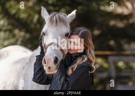 Junge Frau lehnt sich an weißes arabisches Pferd, Augen geschlossen, unscharfer Baumhintergrund, Nahaufnahme Stockfoto