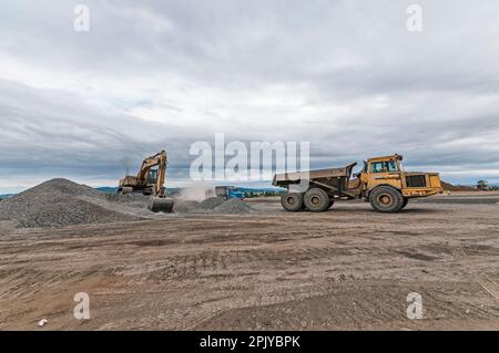 Ein Bagger zieht Steine und Schmutz in der Nähe eines Kipplasters auf einer aktiven Deponie hoch. Stockfoto