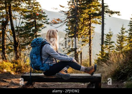 Entspannung beim Wandern in den Bergen. Frau mit Rucksack, die auf einer Holzbank im Wald ruht. Passender Ort zum Campen in den Bergen Stockfoto