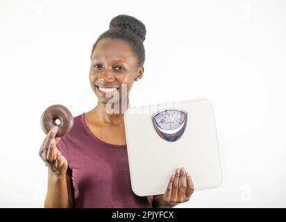 Junge Frau, die zwischen Gewichtsskala und Junk Food auf weißem Hintergrund wählt Stockfoto