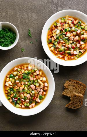 Kalte Okroshka-Suppe mit Wurst, Gemüse und Kvass in Schüsseln auf braunem Hintergrund. Draufsicht, flach liegend Stockfoto