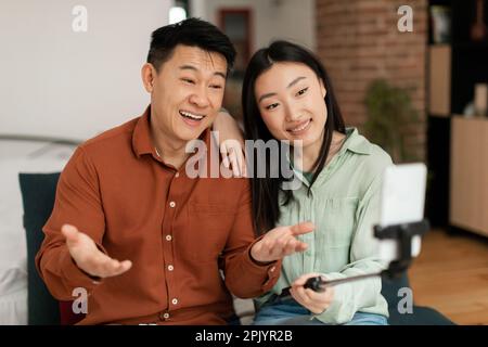 Lächelnde koreanische Ehepartner mit fröhlichen Gesichtern, die auf die Webkamera des Mobiltelefons schauen, Blogger, die Videoblog aufzeichnen Stockfoto