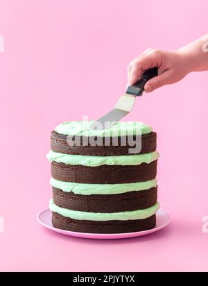 Die Frau macht einen Minzschokoladenkuchen, minimalistisch auf pinkfarbenem Hintergrund. Köstlicher hausgemachter Kuchen mit dunkler Schokolade und Buttercreme mit Minzgeschmack Stockfoto