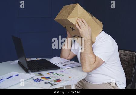 Ein Mann ist ein Geschäftsmann in einem weißen Hemd mit einer Papiertüte auf dem Kopf, mit einem traurigen Emoticon, arbeitet an einem Computer mit Dokumenten, hält seinen Kopf. E Stockfoto