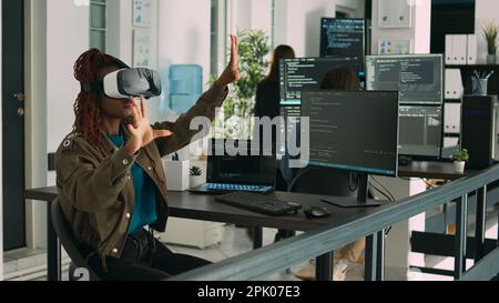 afroamerikanischer Programmierer, der mit VR-Brillen im IT-Entwicklungsbüro arbeitet und ein Virtual-Reality-Headset zur Entwicklung eines Sicherheitsprogramms verwendet. Big Data Admin codiert HTML-Skript mit Datenbank. Stockfoto