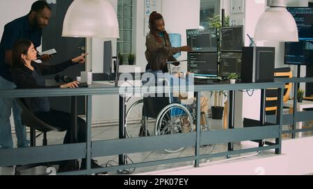 Asiatischer Programmierer mit körperlicher Behinderung arbeitet mit einem afroamerikanischen Entwickler zusammen, um eine Sicherheits-Firewall auf dem Terminalfenster zu erstellen. IT-Ingenieur im Rollstuhlprogrammieren HTML-Skriptcode. Stockfoto