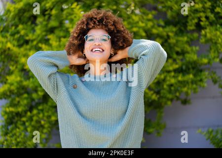 Porträt einer birassischen, fröhlichen jungen Frau mit Händen im Afrohaar, die lächelt und im Hof steht Stockfoto