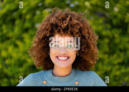 Nahaufnahme einer birassischen, schönen jungen Frau mit Afrohaar, die gegen Pflanzen im Hof lächelt Stockfoto