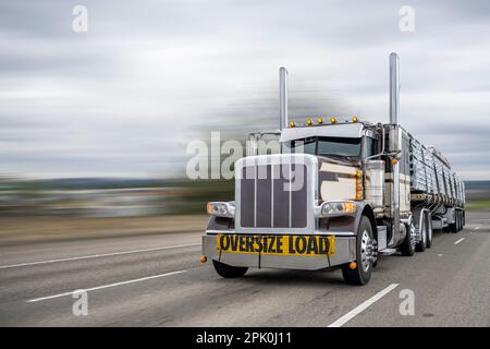 Klassischer beiger Sattelschlepper mit großem Sattelanhänger, mit Schild für Übergröße am Stoßfänger und eingeschaltetem Leichtgewicht für den Transport von Fracht auf Flachbettsattelanhänger Stockfoto