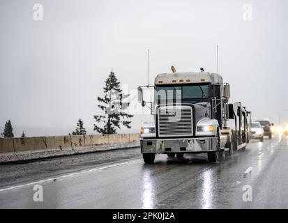 Klassischer schwarzer Lkw-Transporter für industrielle Sattelzugmaschinen mit eingeschaltetem Licht transportiert leere Sattelanhänger auf dem abendlichen regnerischen Hang Stockfoto