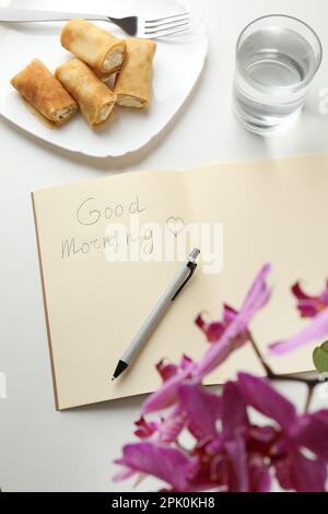 Notizbuch mit Inschrift Good Morning, gefüllte Crepes und ein Glas Wasser neben einer wunderschönen blühenden Orchidee auf einem weißen Tisch, über der Aussicht Stockfoto