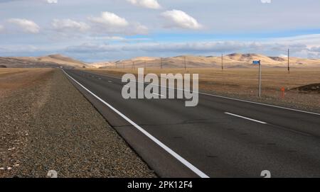 Eine neue zweispurige gepflasterte Straße mit Markierungen überquert die Herbststeppe in Richtung hoher Hügel unter einem bewölkten Himmel. Chuisky-Trakt, Altai, Sibirien, Russi Stockfoto