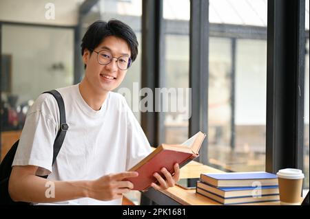 Ein kluger und gutaussehender junger asiatischer Student mit Brille sitzt in einem Café mit Schulbüchern. Stockfoto