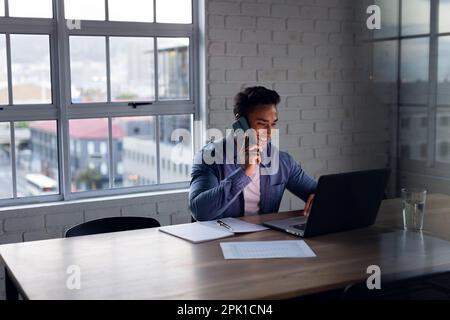 Glücklicher birassistischer Geschäftsmann am Schreibtisch, der ein Notebook benutzt, über ein Smartphone spricht und bis spät in die Nacht im Büro arbeitet Stockfoto