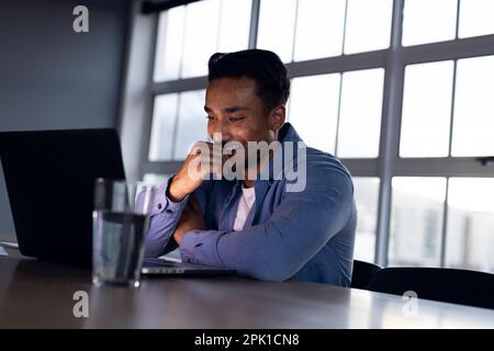 Glücklicher birassistischer Geschäftsmann, der am Schreibtisch sitzt und seinen Laptop benutzt, bis spät in die Nacht im Büro arbeitet Stockfoto