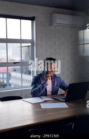 Glücklicher birassistischer Geschäftsmann, der ein Notebook benutzt, über ein Smartphone spricht, bis spät in die Nacht im Büro arbeitet, Kopierraum Stockfoto