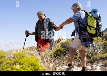 Glückliches älteres, birassisches Paar mit Rucksäcken, Händchen halten und mit Wanderstöcken wandern Stockfoto