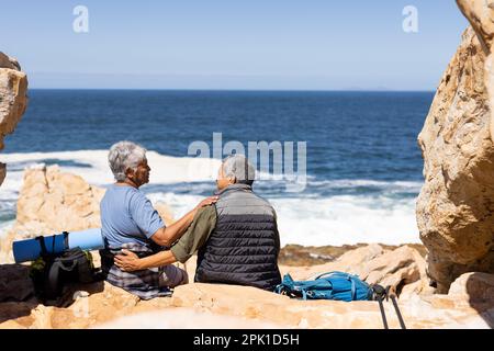Glückliches älteres birassisches Paar, das auf Felsen am Meer sitzt, umarmt und lächelt Stockfoto