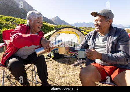 Glückliches älteres birassisches Paar, das auf Campingstühlen sitzt und Kaffee trinkt Stockfoto