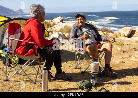 Glückliches älteres birassisches Paar, das auf Campingstühlen sitzt und Kaffee trinkt Stockfoto