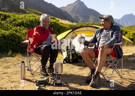 Glückliches älteres birassisches Paar, das auf Campingstühlen sitzt und Kaffee trinkt Stockfoto