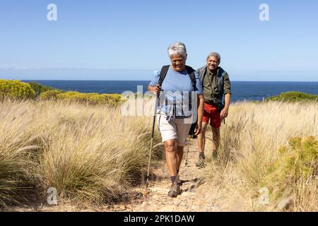 Glückliches älteres birassisches Paar mit Rucksäcken, Wandern mit Wanderstöcken Stockfoto