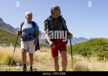 Glückliches älteres birassisches Paar mit Rucksäcken, Wandern mit Wanderstöcken Stockfoto