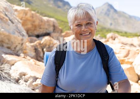 Porträt einer glücklichen älteren, birassischen Frau, die einen Rucksack trägt, in die Kamera schaut und in den Bergen lächelt Stockfoto