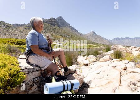 Glückliche ältere, birassische Frau, die in den Bergen wandert, auf einem Felsen sitzt Stockfoto