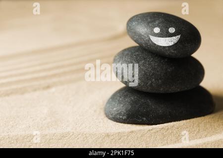 Steine mit gezeichnetem fröhlichem Gesicht auf Sand, Platz für Text. Sei in Harmonie und genieße dein Leben Stockfoto