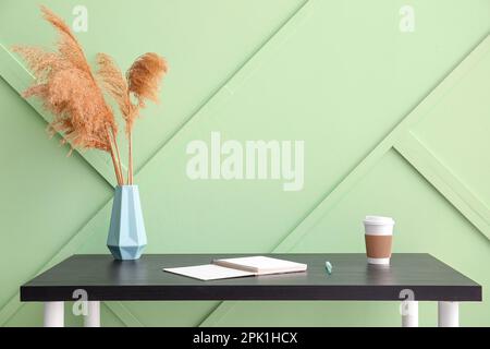 Arbeitsplatz mit Notizbuch, Pampasgras in Vase und Tasse Kaffee an grüner Wand Stockfoto