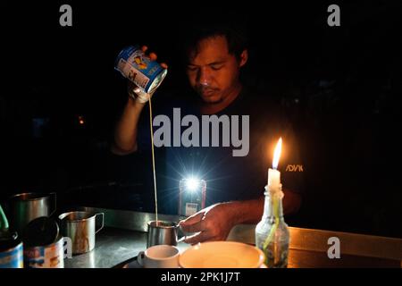 Ein Verkäufer macht Tee bei Kerzenschein während eines Stromausfalls in einem Laden auf der Straße in Yangon. Aufgrund der steigenden Kosten und des Mangels an Wasserkraft während der Trockenzeit rationiert die Militärjunta (Tatmadaw) täglich Strom im gesamten Yangon, wodurch große Teile der Stadt (Townships) täglich komplett geschwärzt wurden. Das tägliche Leben während des tödlichen Bürgerkriegs in Myanmar. Am 1. Februar 2021 ergriff die Militärjunta-Regierung (Tatmadaw) die Macht durch Putsch, sperrte die demokratisch gewählte Regierung ein und stürzte das Land in eine anhaltende humanitäre Krise. Stockfoto