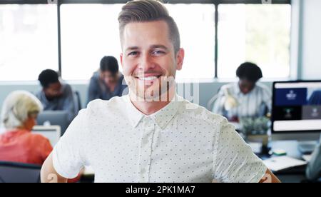 Sei dein Bestes und du wirst der Beste sein. Porträt eines selbstbewussten jungen Geschäftsmannes, der in einem modernen Büro mit seinen Kollegen im Hintergrund arbeitet. Stockfoto