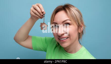 Nahaufnahme eines fröhlichen, süßen blonden Mädchens in einem lässigen Look, das mit dem Kauf eines Hauses auf blauem Hintergrund angibt Stockfoto