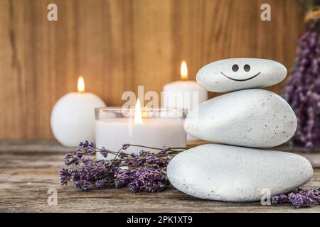 Ein Haufen Steine mit gemaltem Gesicht, Lavendelblumen und Kerzen auf einem Holztisch. Sei in Harmonie und genieße dein Leben Stockfoto