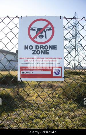 Ein Schild mit der Aufschrift „Keine Drohnenzone“, das an einen hohen Metallzaun gebunden ist. Mit einem Strompylon und Gebäuden im Hintergrund. Stockfoto