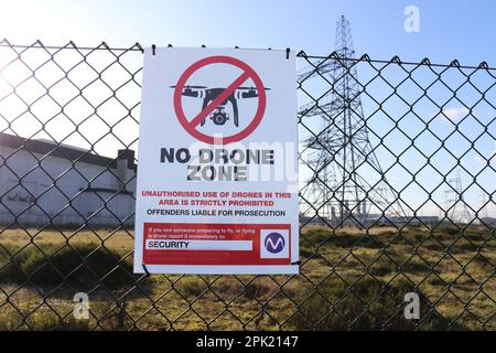 Ein Schild mit der Aufschrift „Keine Drohnenzone“, das an einen hohen Metallzaun gebunden ist. Mit einem Strompylon und Gebäuden im Hintergrund. Stockfoto