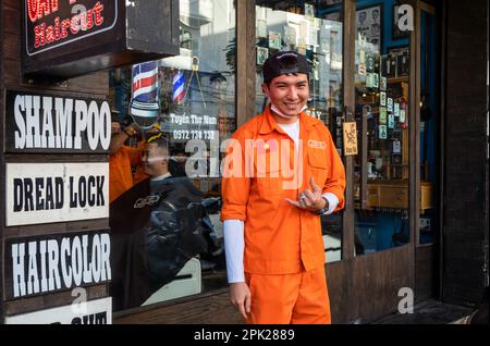 Ein junger Friseur in orangefarbener Uniform steht vor dem Friseursalon, wo er in Pleiku im zentralen Hochland Vietnams arbeitet. Stockfoto