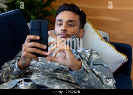 Ein birassischer Soldat, der Uniform trägt, auf dem Sofa mit dem Smartphone liegt Stockfoto