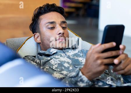 Ein birassischer Soldat, der Uniform trägt, auf dem Sofa mit dem Smartphone liegt Stockfoto