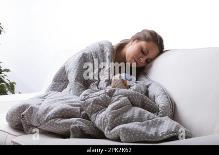 Eine kranke und kalte junge Frau sucht Trost. Sie sitzt in einer warmen Decke auf einer weißen Couch und bläst auf das dampfende heiße Getränk in der Tasse, die sie hält Stockfoto