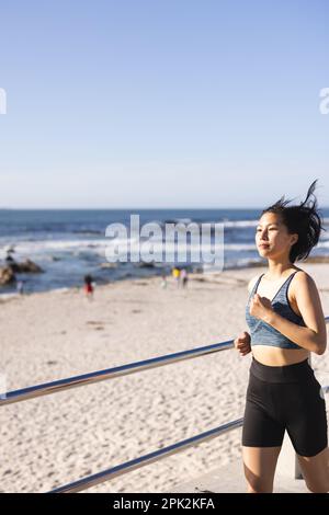 Eine glückliche asiatische Frau, die Sportbekleidung trägt und auf der Promenade am Meer läuft Stockfoto