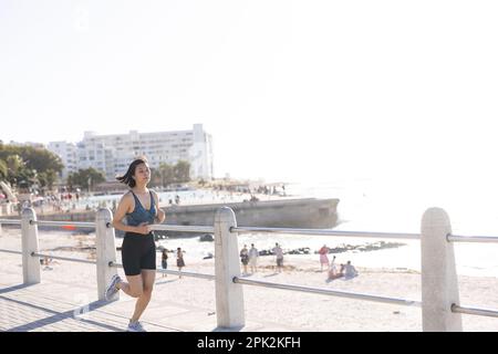 Eine glückliche asiatische Frau, die Sportbekleidung trägt und auf der Promenade am Meer läuft Stockfoto