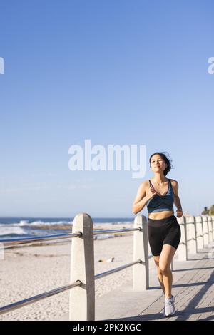 Eine glückliche asiatische Frau, die Sportbekleidung trägt und auf der Promenade am Meer läuft Stockfoto