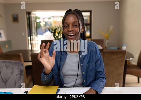 Glückliche afroamerikanische Teenagerin, die am Tisch sitzt und Videoanrufe hat Stockfoto